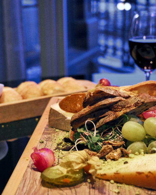 A board with cheese, bread and grapes next to a glass of red wine